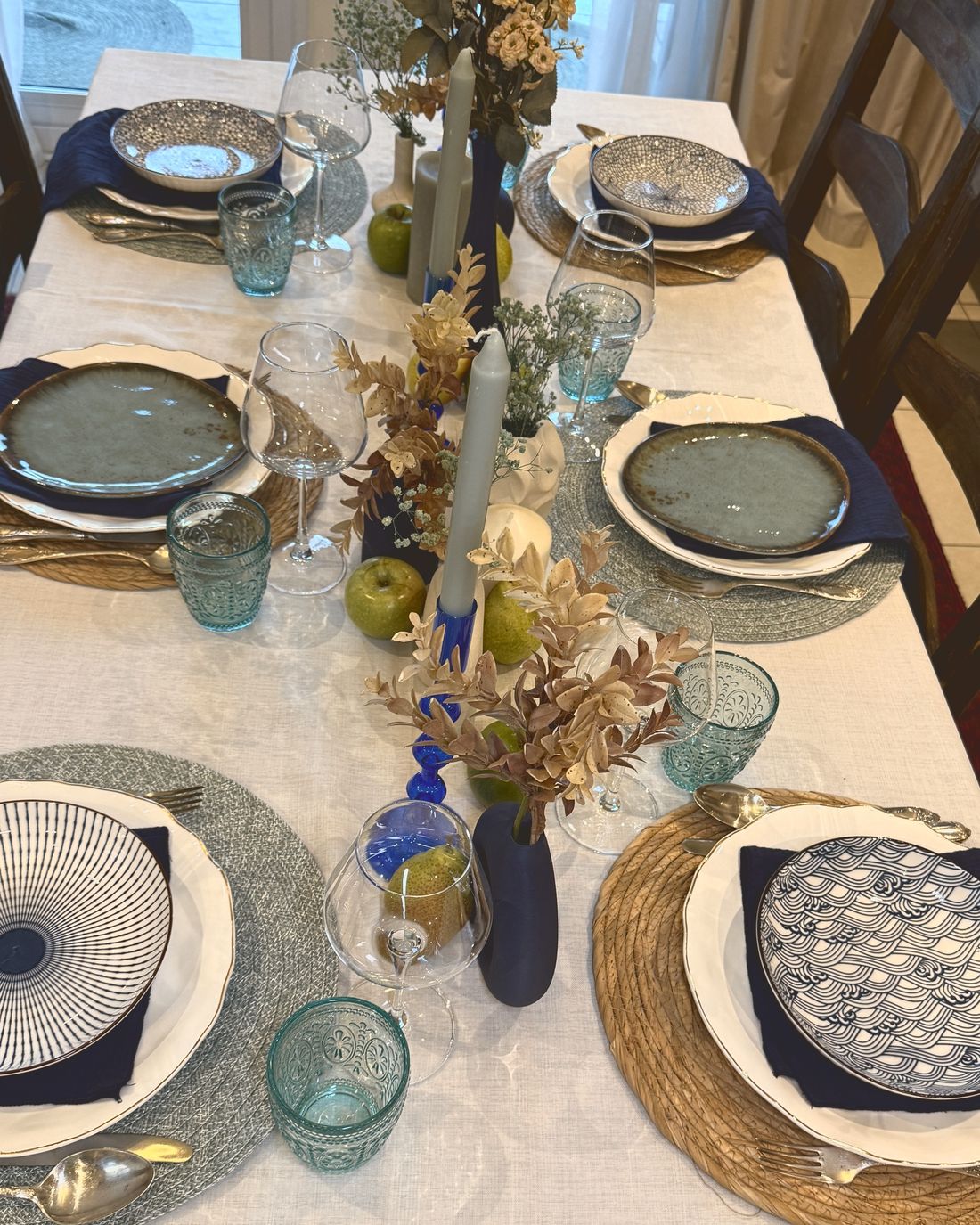 A full dinner table with blue and teal plates, glasses and dried flowers